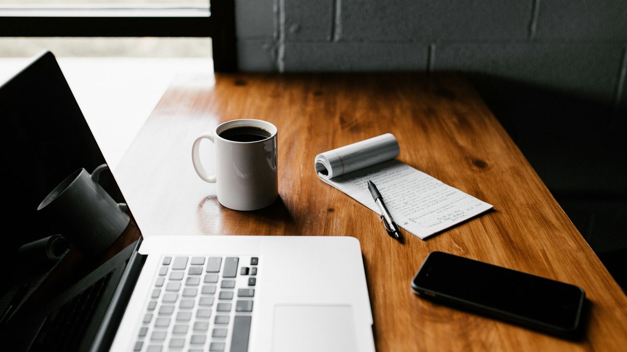  Das Bild zeigt einen minimalistischen Arbeitsplatz. Auf einem Holztisch befinden sich ein MacBook Pro, ein weißer Keramikbecher und ein schwarzes Smartphone. Der Hintergrund ist unscharf, wodurch der Fokus auf den Gegenständen auf dem Tisch liegt.