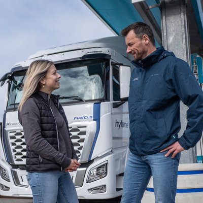 Ein Mann und eine Frau unterhalten sich vor einem modernen Wasserstoff-Lkw an einer Tankstelle. Beide tragen Freizeitkleidung, wobei der Mann eine blaue Jacke und die Frau eine schwarze Jacke trägt.