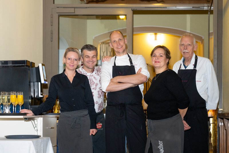 Das Bild zeigt fünf Personen, die als Team eines Restaurants oder einer Gaststätte zusammenarbeiten. Sie tragen teils Arbeitskleidung wie Schürzen und lächeln in einer einladenden und freundlichen Atmosphäre in die Kamera, während im Hintergrund ein Eingangsbereich mit Holzelementen zu sehen ist.