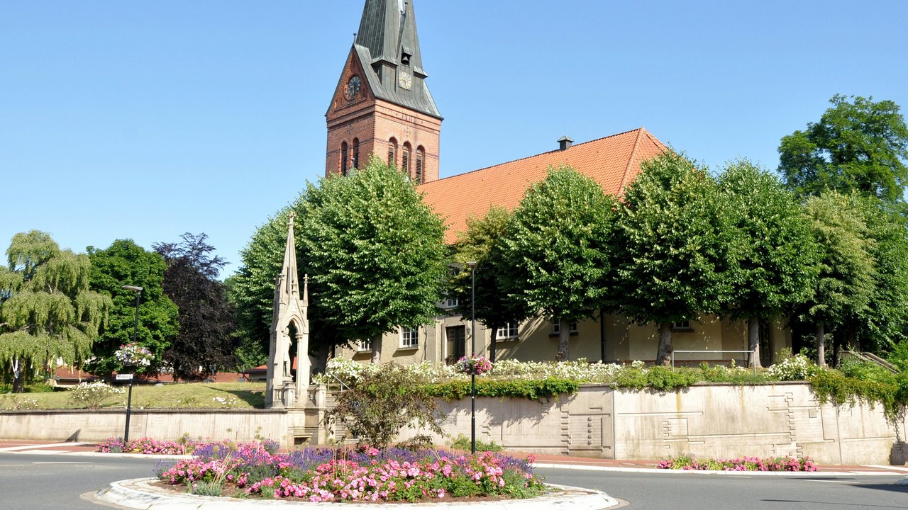  Das Bild zeigt eine malerische Kirche mit einem markanten Glockenturm, eingebettet in eine üppige, parkähnliche Umgebung. Der Kirchplatz ist mit einer Blumenrabatte und Brunnen geschmückt, was der Szene eine idyllische und einladende Atmosphäre verleiht.