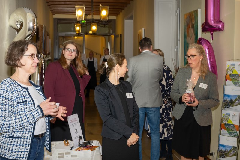 Das Bild zeigt eine Gruppe von Menschen, die in einem Flur stehen und miteinander interagieren, während einige lächeln und sich unterhalten. Im Vordergrund sind drei Frauen zu sehen, die an einem Tisch stehen, während im Hintergrund weitere Gäste an einer Veranstaltung teilnehmen.