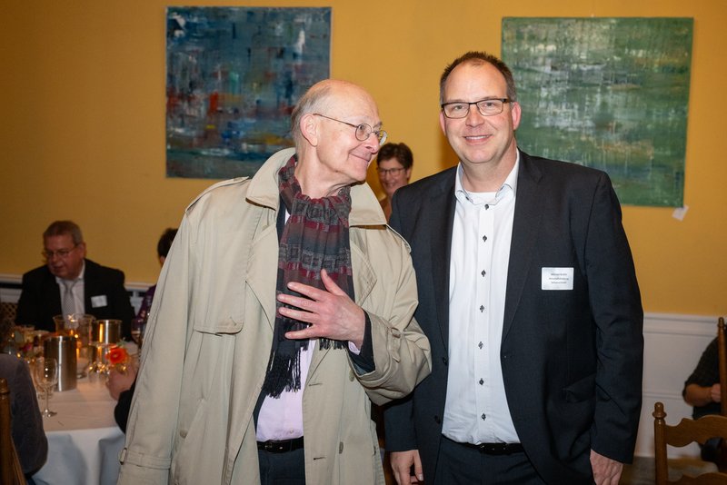 Das Bild zeigt zwei Männer in einem festlichen Raum mit dekorierten Tischen im Hintergrund. Der Mann links trägt einen hellen Mantel und hat einen Schal um, während der Mann rechts in einem dunklen Anzug gekleidet ist. Beide lächeln und scheinen sich in einer geselligen Atmosphäre zu befinden. Im Hintergrund sind weitere Personen und Kunstwerke an den Wänden zu sehen.