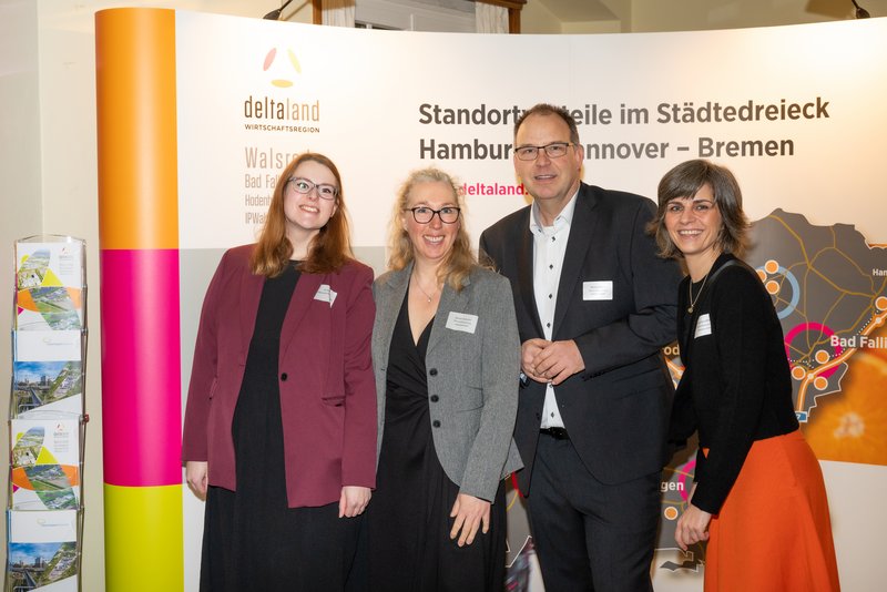 Das Bild zeigt eine Gruppe von vier Personen, die vor einem farbenfrohen Hintergrund mit Informationen über die Region "deltaland" stehen. Alle Teilnehmer lächeln und tragen Namensschilder, was auf eine Veranstaltung oder Präsentation hinweist.