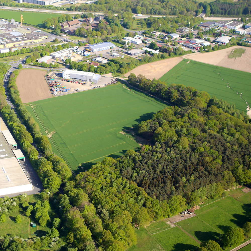 Erweiterungsflächen in Bad Fallingbostel | Ⓒ Deltaland | Erstellt von: GÖ-Flug Das Bild zeigt eine Luftaufnahme einer ländlich-industriellen Gegend mit weitläufigen Feldern, einem Waldstück und mehreren Fabrik- sowie Lagergebäuden. Am oberen Bildrand sind Wohn- und Gewerbegebiete zu erkennen, die von grünen Landschaftsflächen umgeben sind.