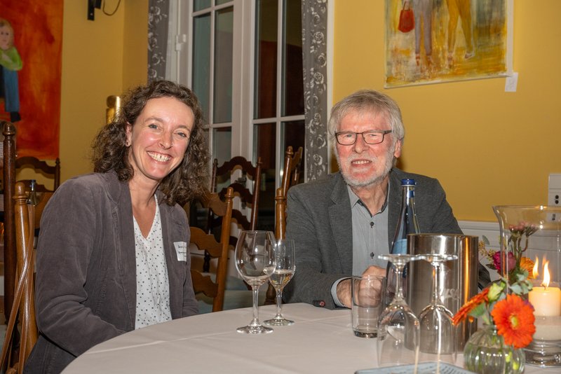 Das Bild zeigt eine Frau und einen Mann, die an einem Tisch sitzen und lächeln. Beide wirken entspannt und freundlich. Auf dem Tisch sind mehrere Gläser, eine Flasche und eine kleine Blumendekoration zu sehen. Im Hintergrund sind einige Gemälde an der Wand zu erkennen.