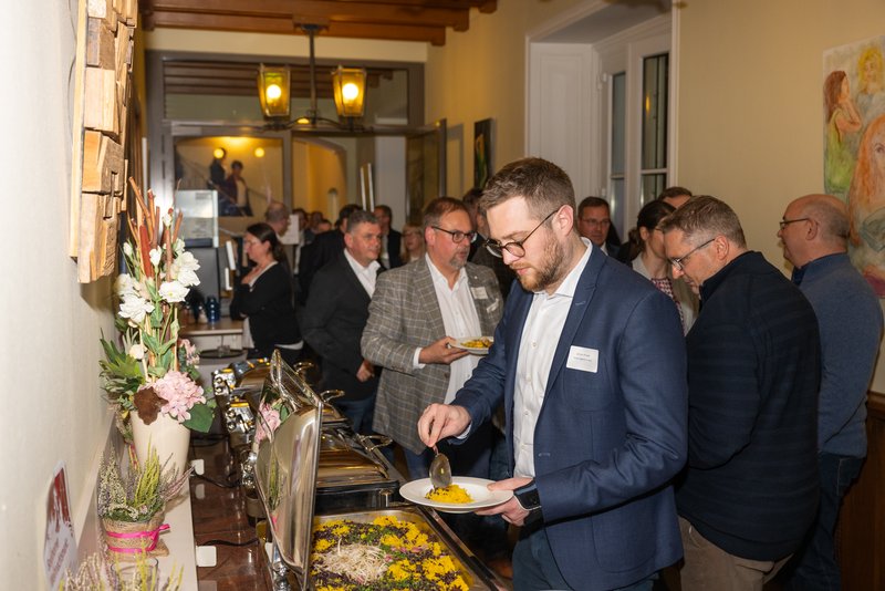 Das Bild zeigt eine Gruppe von Menschen, die an einem Buffet in einem hellen Raum stehen. Ein Mann im Vordergrund füllt sich gerade einen Teller mit Essen, während andere Gäste im Hintergrund miteinander sprechen und sich ebenfalls bedienen.