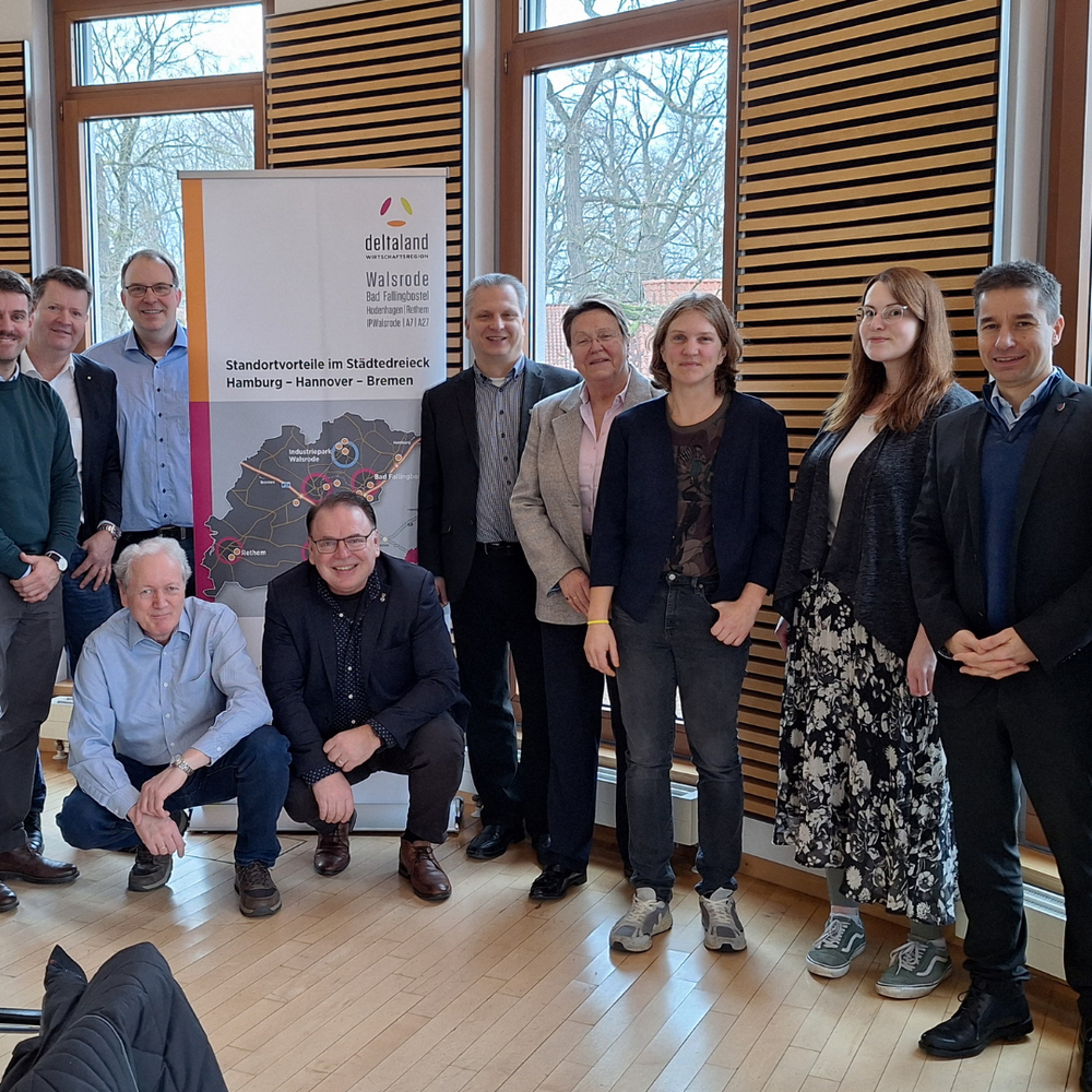 | Ⓒ Deltaland In einem hellen Raum mit großen Fenstern posiert eine Gruppe von 15 Personen für ein Gruppenfoto vor einem Banner. Die Anwesenden stehen und sitzen lächelnd zusammen.