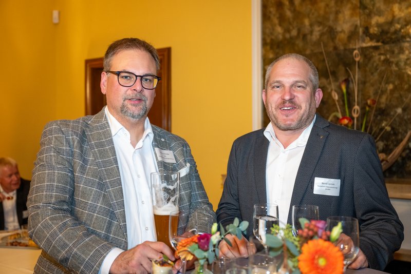 Das Bild zeigt zwei Personen, die an einem Stehtisch stehen und lächeln, während sie Getränke und Snacks vor sich haben. Die Umgebung wirkt festlich und einladend, was auf eine gesellige Veranstaltung hindeutet.