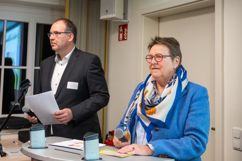 Das Bild zeigt zwei Personen, die an einem Tisch stehen, während sie ans Publikum sprechen. Der Mann auf der linken Seite trägt einen dunklen Anzug und hält ein Blatt Papier in der Hand. Die Frau auf der rechten Seite trägt einen blauen Blazer und einen bunten Schal. Beide wirken konzentriert und freundlich. Im Hintergrund sind Fenster und einige technische Geräte zu sehen, was auf eine formelle Veranstaltung oder Präsentation hindeutet. Die Atmosphäre ist professionell und engagiert.