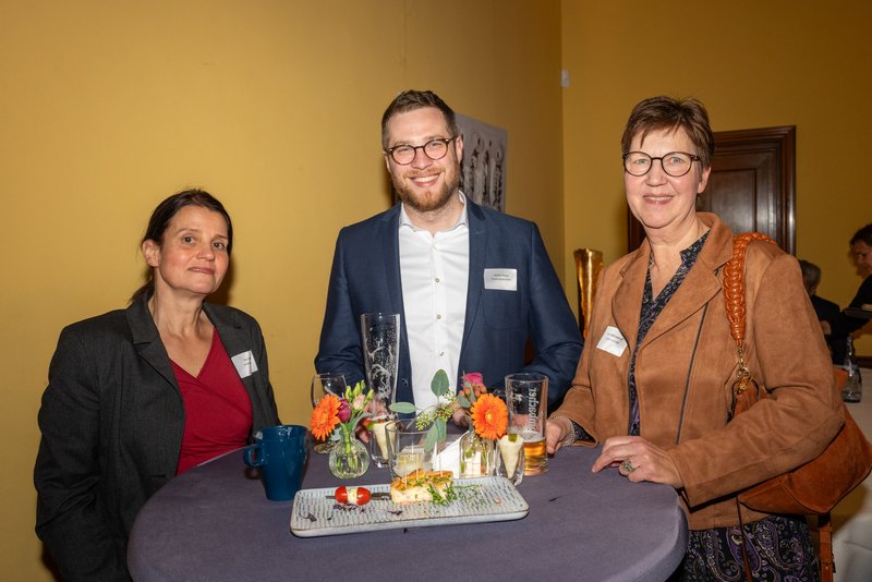 Das Bild zeigt drei Personen, die an einem Stehtisch stehen und lächeln, während sie Getränke und Snacks vor sich haben. Die Umgebung wirkt festlich und einladend, was auf eine gesellige Veranstaltung hindeutet.