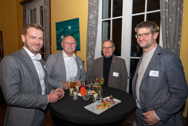 Das Bild zeigt vier Personen, die an einem Stehtisch stehen und lächeln, während sie Getränke und Snacks vor sich haben. Die Umgebung wirkt festlich und einladend, was auf eine gesellige Veranstaltung hindeutet.