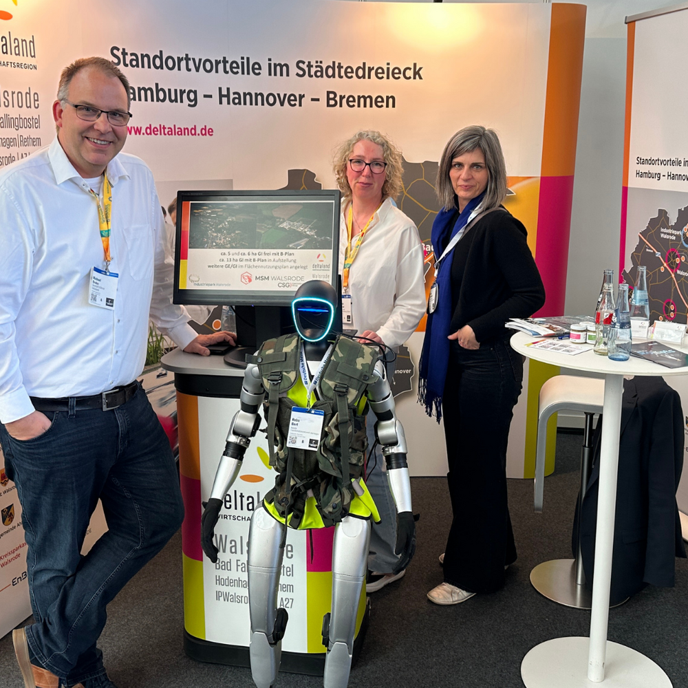 Standteam mit Roboter | Ⓒ Deltaland Am Deltaland-Messestand stehen ein Mann im weißen Hemd (links), eine Frau mit Brille in weißer Bluse (Mitte) und eine Frau in dunkler Jacke (rechts). Vor ihnen befindet sich ein humanoider Roboter mit Deltaland-Branding auf der Körpervorderseite. Im Hintergrund ist das Roll-up-Banner mit dem Slogan „Standortvorteile im Städtedreieck Hamburg – Hannover – Bremen" sowie ein Prospektständer mit Informationsmaterial sichtbar.