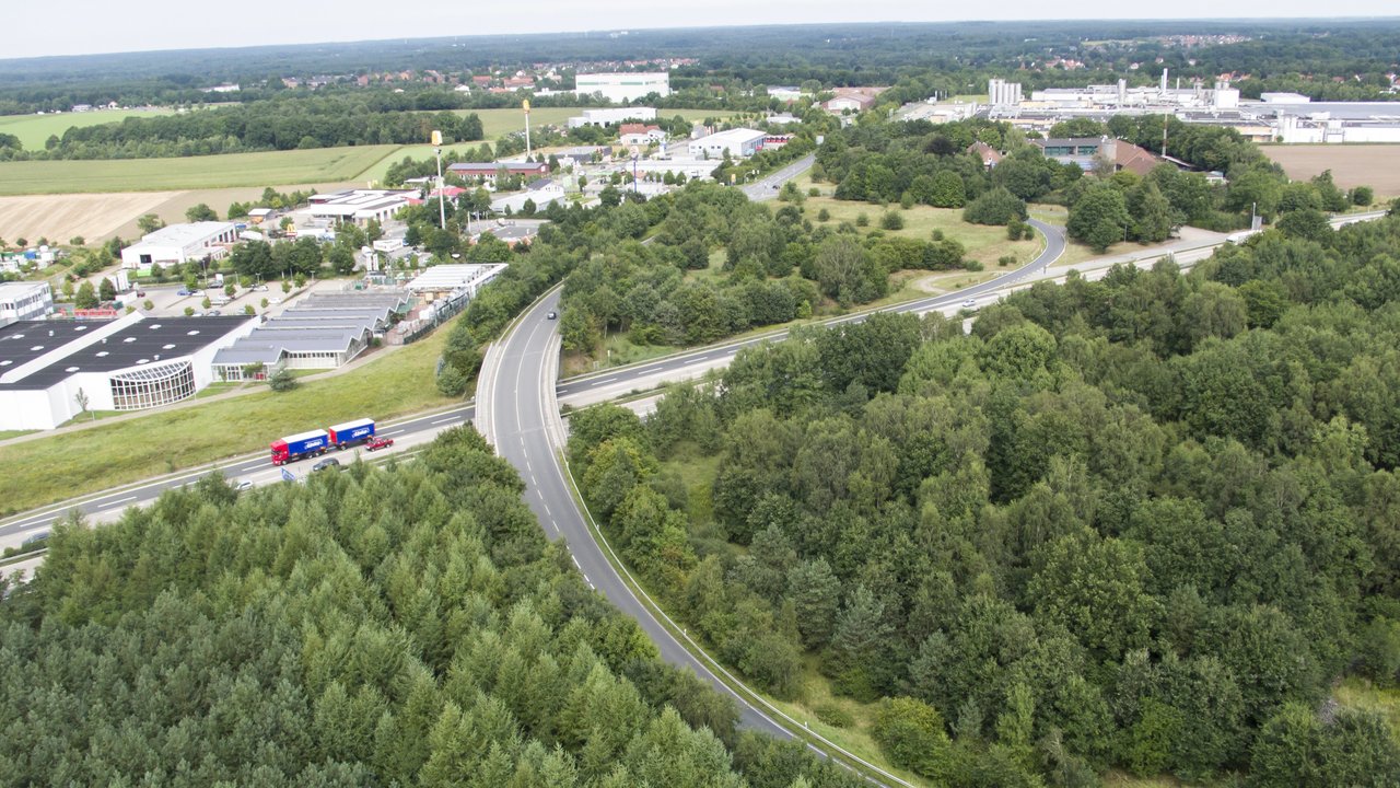  Luftaufnahme des A27parks Walsrode, einem Industrie- und Gewerbegebiet, umgeben von üppiger grüner Vegetation. Im Vordergrund sind Straßen zu sehen, auf denen Fahrzeuge fahren. Im Hintergrund sind Gebäude und Fabrikanlagen sichtbar