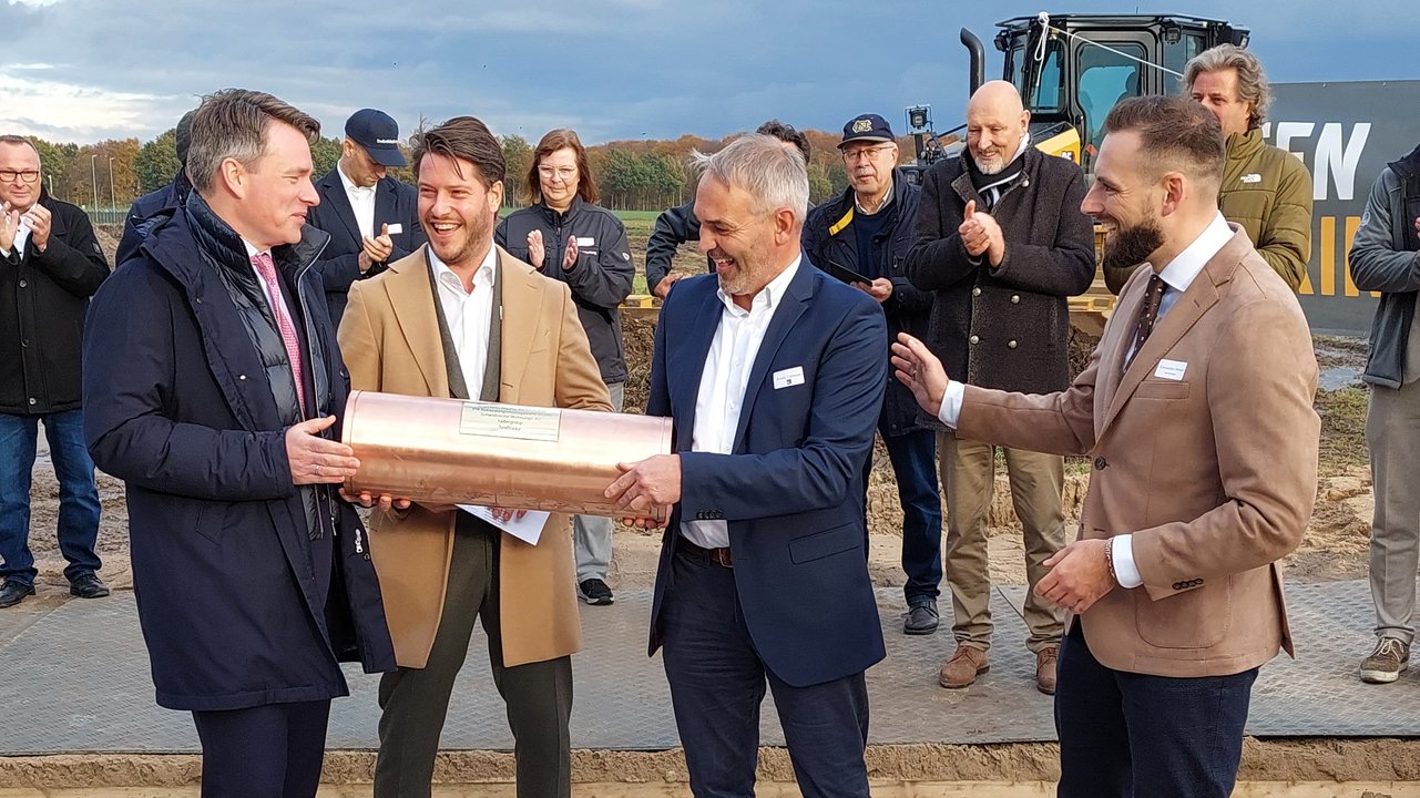 Das Bild zeigt eine Gruppe von Männern, die bei einer Grundsteinlegung eine kupferne Zeitkapsel halten. Im Hintergrund sind Zuschauer und ein Bagger zu sehen, während die Männer vorne lächeln und sich auf die Zeremonie konzentrieren.