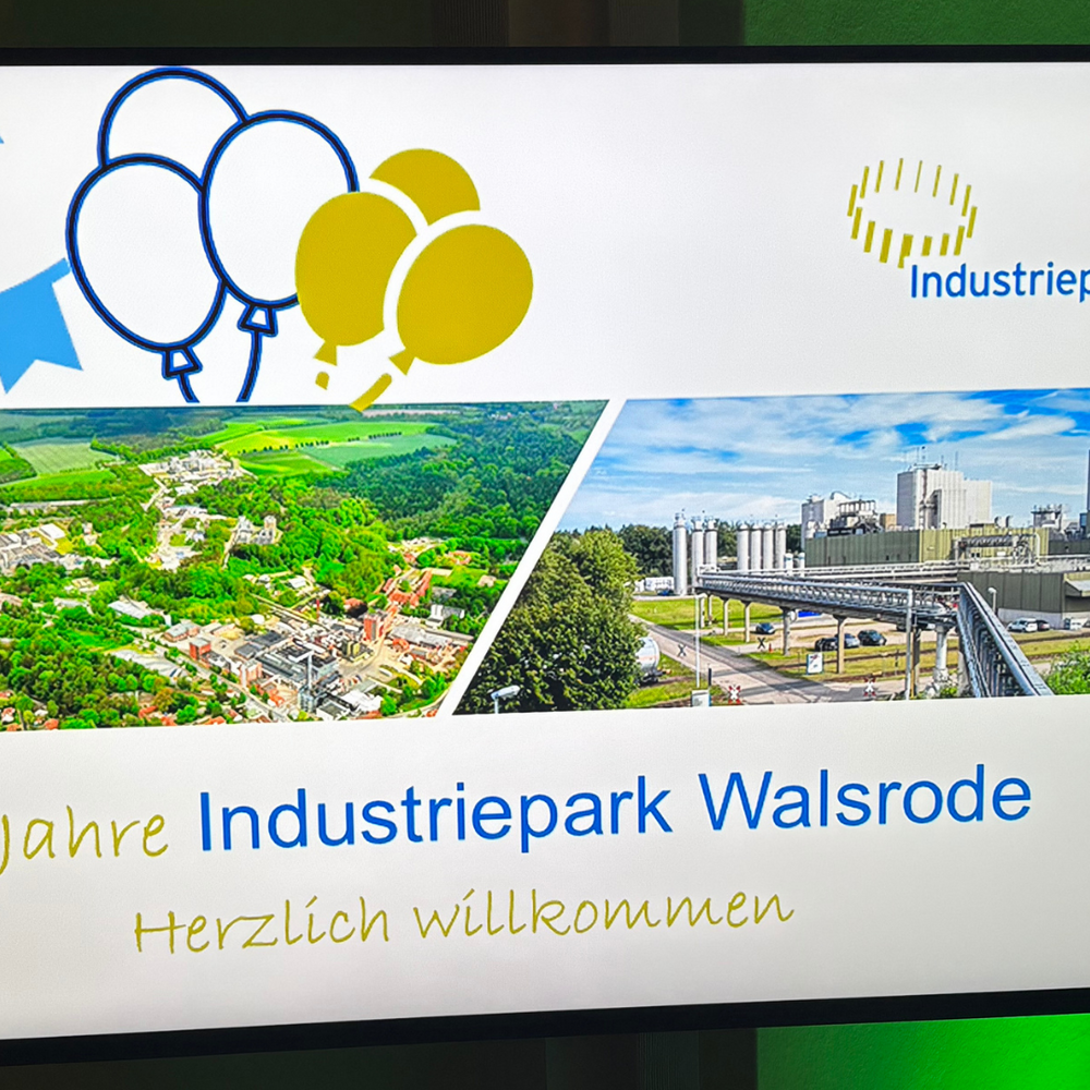 | Ⓒ Deltaland Bildschirm mit der Willkommensfolie „25 Jahre Industriepark Walsrode – Herzlich willkommen”. Die Folie zeigt das Logo des Industrieparks, Luftaufnahmen des Geländes sowie Wimpelketten und Luftballons in Blau und Gelb.