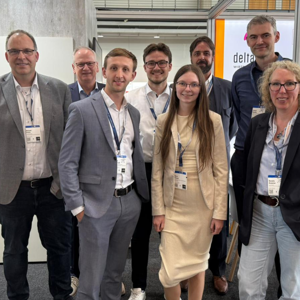 | Ⓒ Deltaland Acht Personen in Business-Kleidung stehen lächelnd für ein Gruppenfoto am deltaland-Messestand, alle tragen Besucherausweise am Lanyard. Im Hintergrund ist das deltaland-Logo auf einer beleuchteten Wand sowie eine Regionalkarte mit Standortmarkierungen zu sehen. Links eine rote Infotafel mit Grundrissplänen, rechts ein Hinweisschild mit der Standnummer „G24".