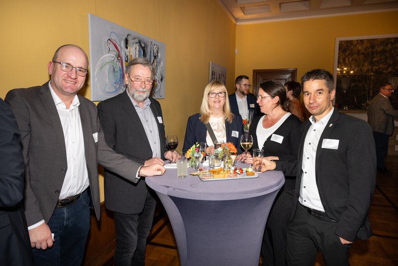 Das Bild zeigt eine Gruppe von Menschen, die an einem Stehtischen stehen. Einige Personen lächeln in die Kamera. Die Anwesenden sind in formeller Kleidung und scheinen an einer Veranstaltung teilzunehmen. Im Hintergrund sind Bilder zu sehen, was auf eine elegante Umgebung hindeutet.