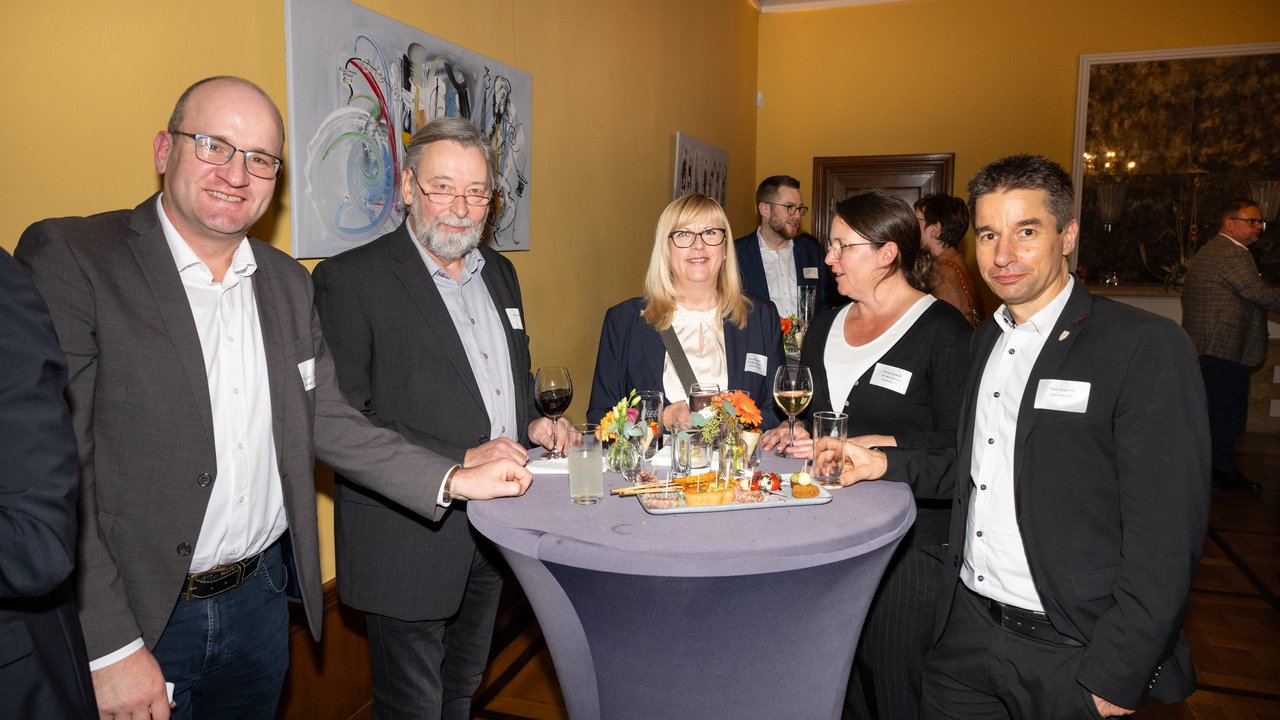 Das Bild zeigt eine Gruppe von Menschen, die an einem Stehtischen stehen. Einige Personen lächeln in die Kamera. Die Anwesenden sind in formeller Kleidung und scheinen an einer Veranstaltung teilzunehmen. Im Hintergrund sind Bilder zu sehen, was auf eine elegante Umgebung hindeutet.