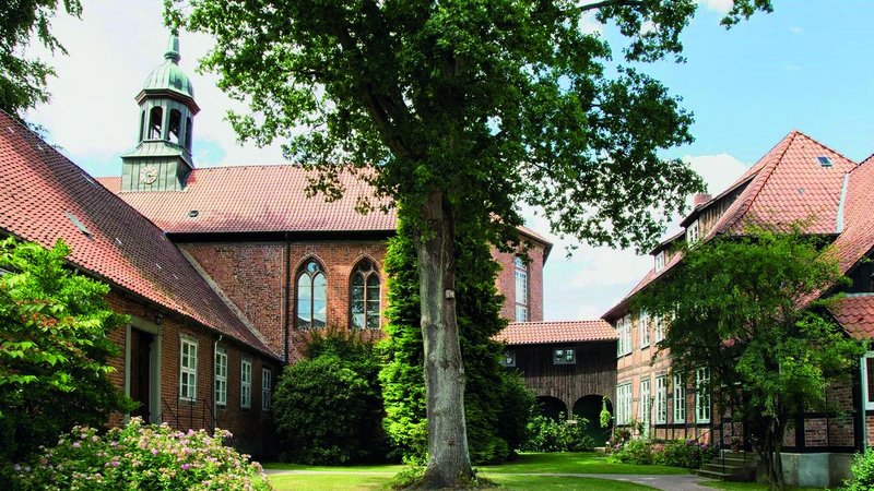 Das Bild zeigt ein malerisches, historisches Gebäudeensemble in einer parkähnlichen Umgebung. Die Architektur mit dem markanten Glockenturm sowie die üppige Vegetation mit den alten Bäumen verleihen der Szene einen idyllischen und geschichtsträchtigen Charakter.