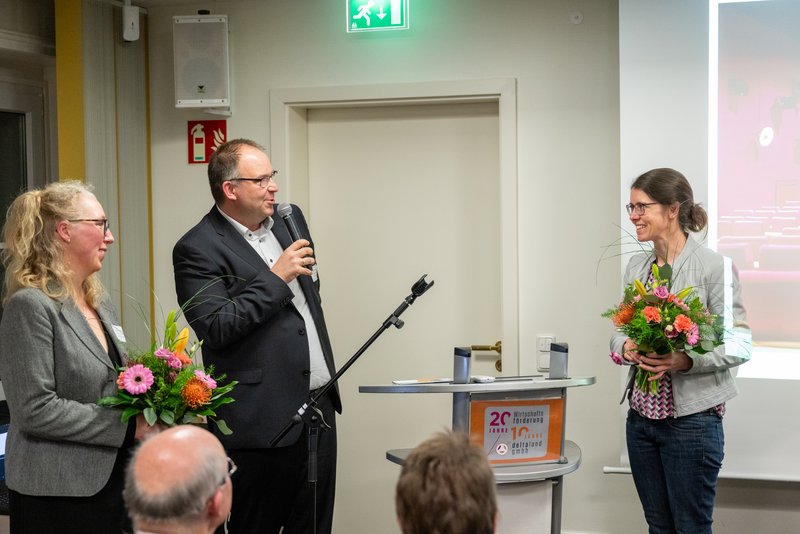 Das Bild zeigt eine kleine Veranstaltung, bei der ein Mann mit Mikrofon zwischen zwei Frauen steht, die jeweils einen Blumenstrauß in der Hand halten. Die Atmosphäre ist feierlich, und im Hintergrund ist eine Präsentation zu sehen, die auf eine Leinwand projiziert wird.