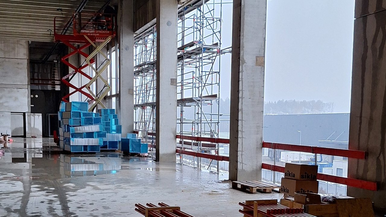 Das Bild zeigt einen Blick in einen großen, industriellen Innenraum. Hier sind zahlreiche Stahlkonstruktionen, Stromkabel und Regalsysteme zu sehen, die auf eine Produktions- oder Lagerhalle hindeuten.