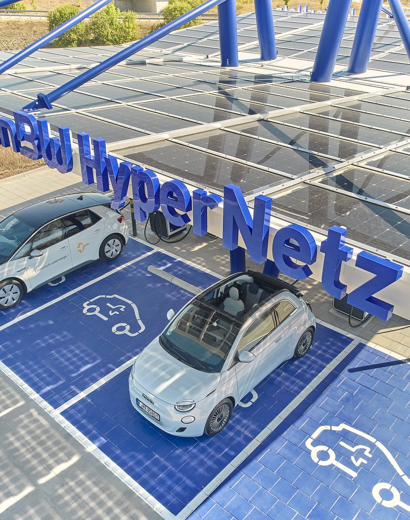 Das Bild zeigt eine große Photovoltaik-Anlage, die ein Parkplatz für Elektrofahrzeuge überdacht. Auf den Solarpaneelen sind zwei weiße Elektroautos geparkt, die offensichtlich an der Ladestation aufgeladen werden. Die Anlage vermittelt einen modernen, nachhaltigen Eindruck und bietet Autofahrern die Möglichkeit, ihre Fahrzeuge umweltfreundlich aufzuladen.