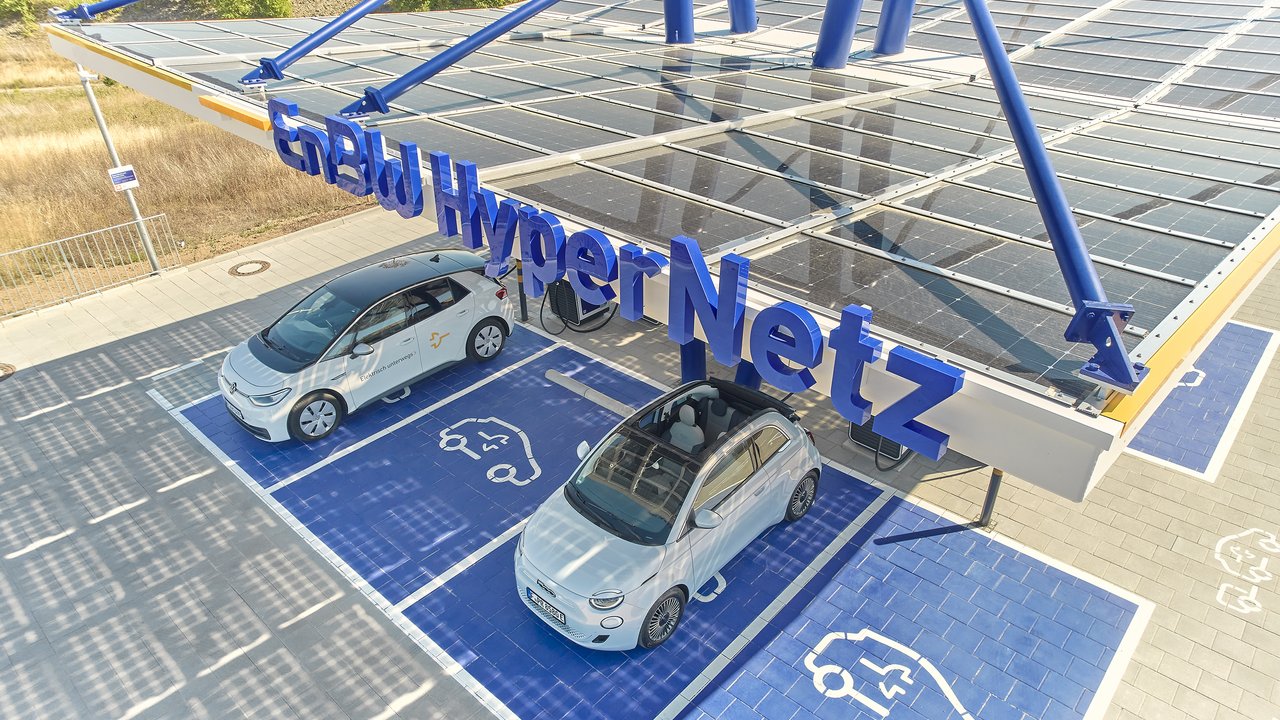 Das Bild zeigt eine große Photovoltaik-Anlage, die ein Parkplatz für Elektrofahrzeuge überdacht. Auf den Solarpaneelen sind zwei weiße Elektroautos geparkt, die offensichtlich an der Ladestation aufgeladen werden. Die Anlage vermittelt einen modernen, nachhaltigen Eindruck und bietet Autofahrern die Möglichkeit, ihre Fahrzeuge umweltfreundlich aufzuladen.