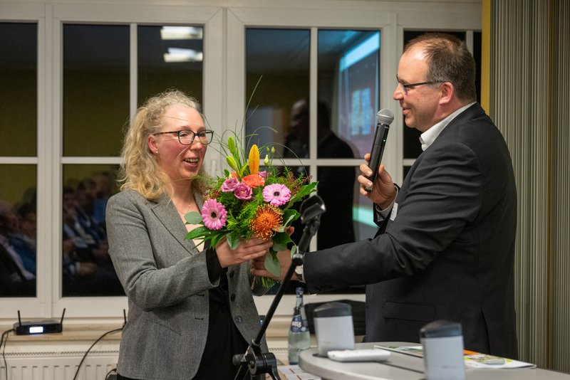 Das Bild zeigt einen Mann, der mit einem Mikrofon in der Hand eine Frau anspricht, während sie einen Blumenstrauß entgegennimmt. 