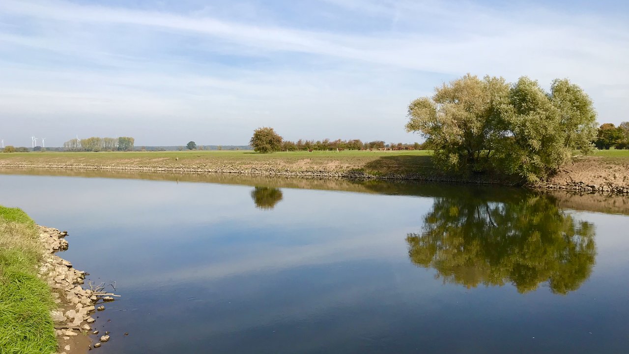  Das Bild zeigt eine idyllische Naturlandschaft mit einem ruhigen See, dessen Oberfläche die umliegenden Bäume und den bewölkten Himmel perfekt widerspiegelt. Die Uferbefestigung aus Steinen und die angrenzenden Wiesenflächen verleihen der Szene einen harmonischen und friedlichen Charakter.