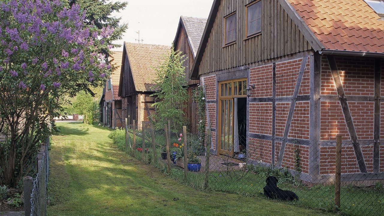  Das Bild zeigt eine malerische ländliche Umgebung mit traditionellen Fachwerkgebäuden, die in eine üppige Grünlandschaft eingebettet sind. Der Kiesweg führt zwischen den liebevoll gestalteten Häusern und Gärten hindurch und vermittelt eine idyllische, friedliche Atmosphäre.