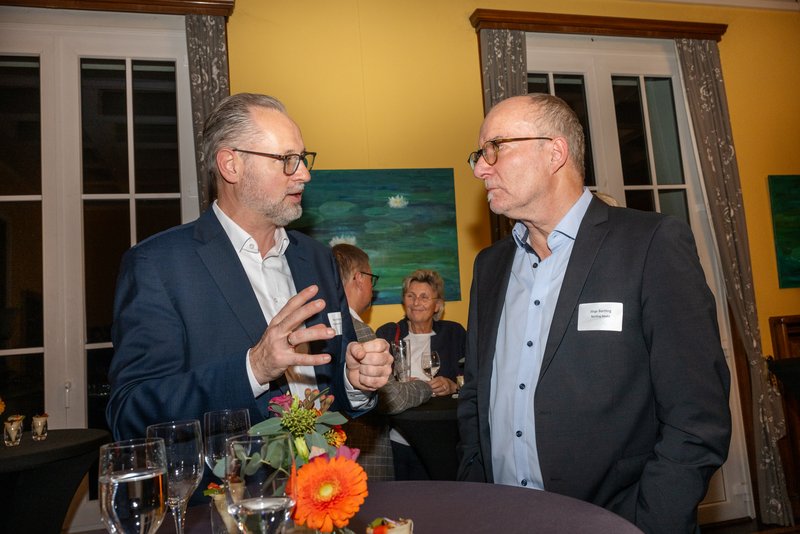 Das Bild zeigt zwei Männer, die an einem Stehtisch stehen und sich angeregt unterhalten. Beide tragen formelle Kleidung; einer hat eine Brille. Auf dem Tisch stehen Gläser und eine kleine Blumendekoration. Im Hintergrund sind weitere Gäste zu sehen, die ebenfalls an Gesprächen teilnehmen.