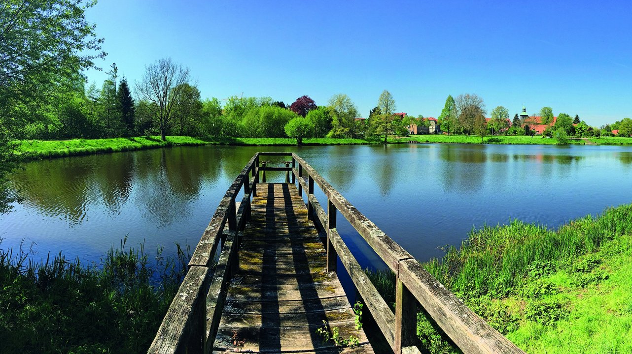  Das Bild zeigt eine idyllische Landschaft mit einem kleinen See, an dessen Ufer sich ein langer, hölzerner Steg erstreckt. Die Umgebung ist von grünen Bäumen und Wiesen gesäumt, die sich in dem ruhigen Wasser des Sees spiegeln, was eine friedliche und natürliche Atmosphäre vermittelt.