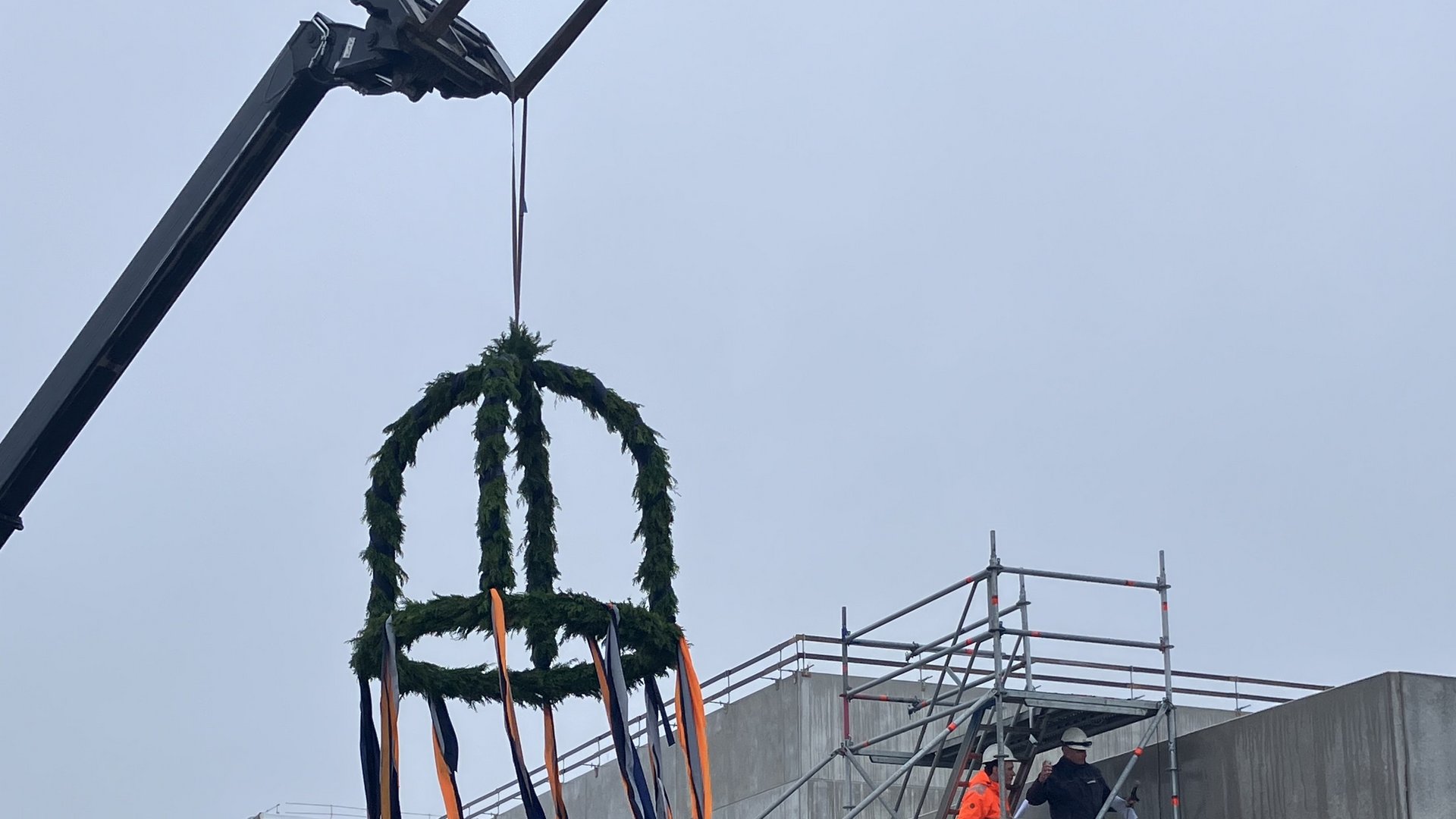 Das Bild zeigt eine Baustelle mit einem Gerüst, an dem Werbebanner für "Ten Brink" angebracht sind. Im Vordergrund befindet sich eine auffällige Skulptur oder Dekoration, die aus Stahlkonstruktionen und einem Kranz aus Grünpflanzen besteht.