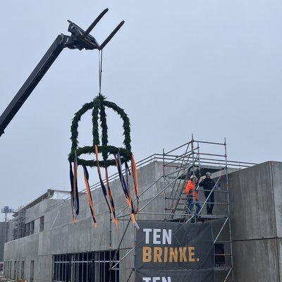 Das Bild zeigt eine Baustelle mit einem Gerüst, an dem Werbebanner für "Ten Brink" angebracht sind. Im Vordergrund befindet sich eine auffällige Skulptur oder Dekoration, die aus Stahlkonstruktionen und einem Kranz aus Grünpflanzen besteht.