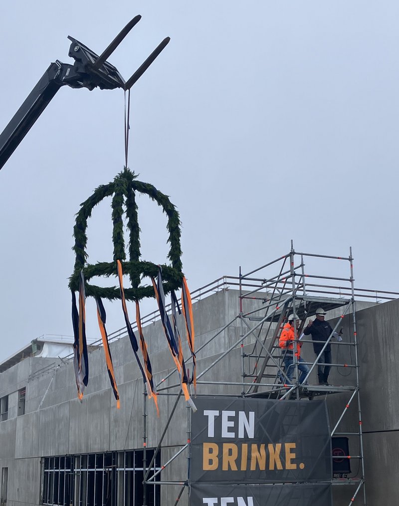 Das Bild zeigt eine Baustelle mit einem Gerüst, an dem Werbebanner für "Ten Brink" angebracht sind. Im Vordergrund befindet sich eine auffällige Skulptur oder Dekoration, die aus Stahlkonstruktionen und einem Kranz aus Grünpflanzen besteht.