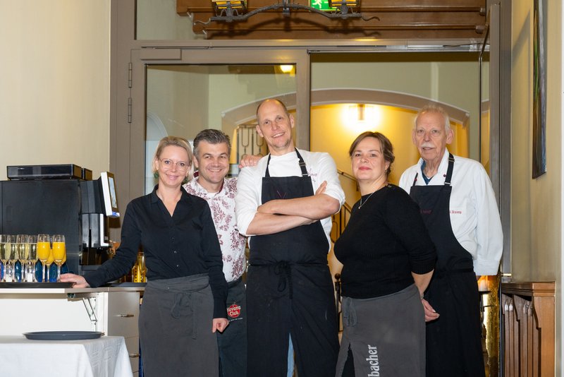 Das Bild zeigt fünf Personen, die als Team eines Restaurants oder einer Gaststätte zusammenarbeiten. Sie tragen teils Arbeitskleidung wie Schürzen und lächeln in einer einladenden und freundlichen Atmosphäre in die Kamera, während im Hintergrund ein Eingangsbereich mit Holzelementen zu sehen ist.