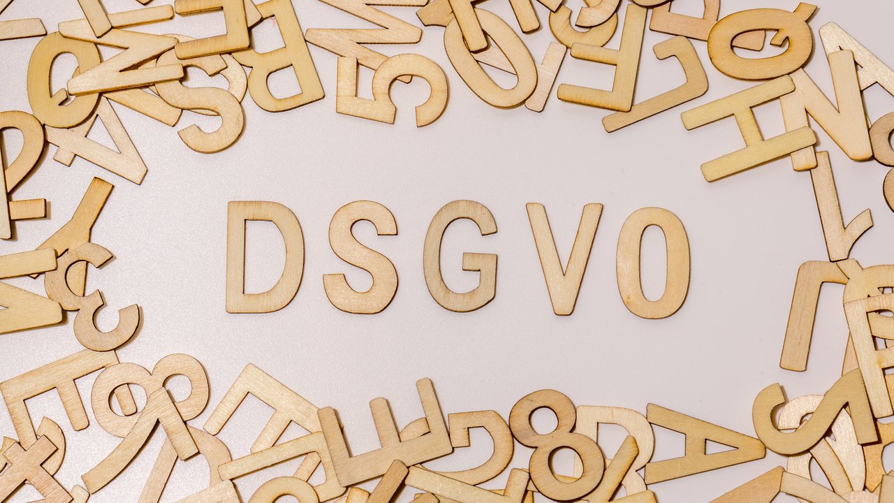  Das Bild zeigt den Begriff "DSGVO" in großen Holzbuchstaben, die auf einer weißen Oberfläche liegen. Rund um die Buchstaben sind viele weitere Holzbuchstaben und -zahlen verstreut. Der Begriff steht für die Datenschutz-Grundverordnung.