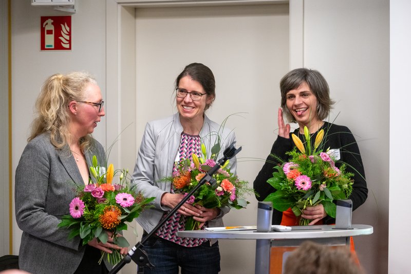 Das Bild zeigt eine Gruppe von drei Personen, die an einem Tisch stehen und lächeln, während sie bunte Blumensträuße in den Händen halten. Die Atmosphäre wirkt festlich und feierlich, was auf eine besondere Veranstaltung oder Ehrung hinweist.