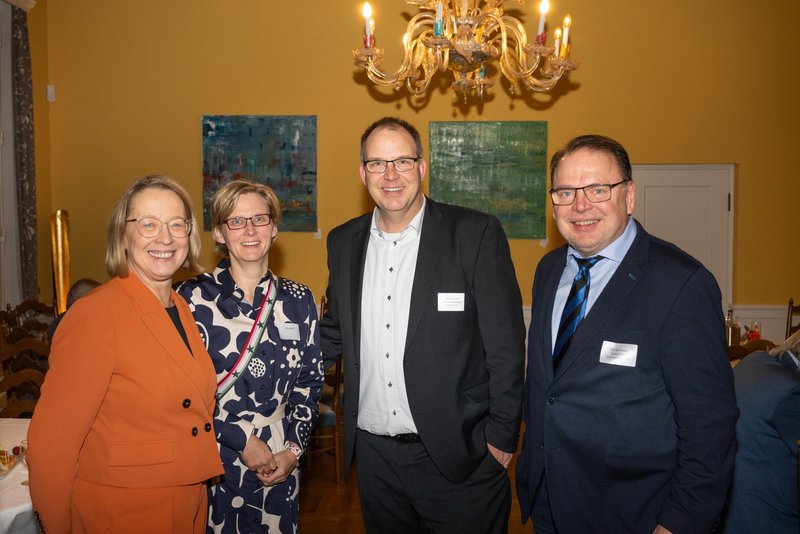 Das Bild zeigt vier Personen in einem festlichen Raum mit einem Kronleuchter. Sie stehen lächelnd zusammen, während sie sich unterhalten. Die Frau links trägt einen orangefarbenen Blazer, die anderen sind in formeller Kleidung. Im Hintergrund sind dekorative Gemälde zu sehen.