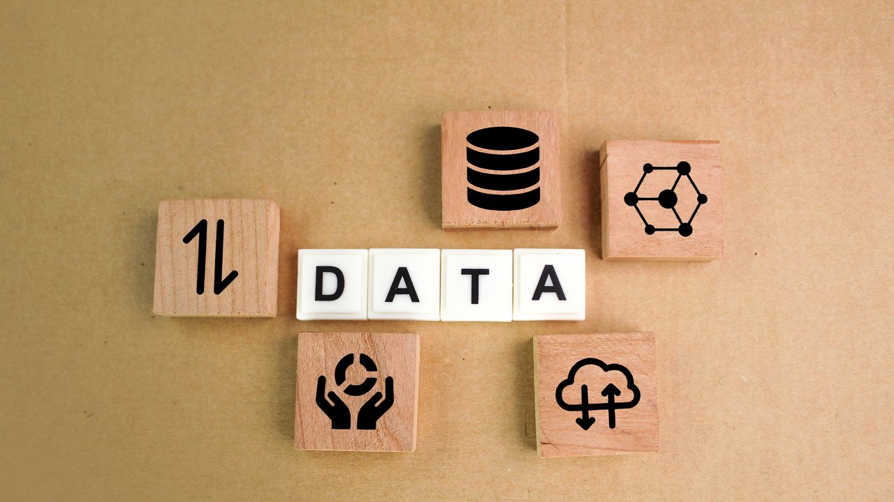  Das Bild zeigt das Wort "DATA", das aus weißen Buchstabenwürfeln besteht, die auf einer beigen Oberfläche angeordnet sind. Um das Wort herum befinden sich sechs Holzblöcke mit verschiedenen Symbolen, die Daten und Technologie darstellen. Diese Symbole umfassen ein Diagramm, eine Datenbank, eine Vernetzung, ein Cloud-Symbol und ein Recycling-Symbol.