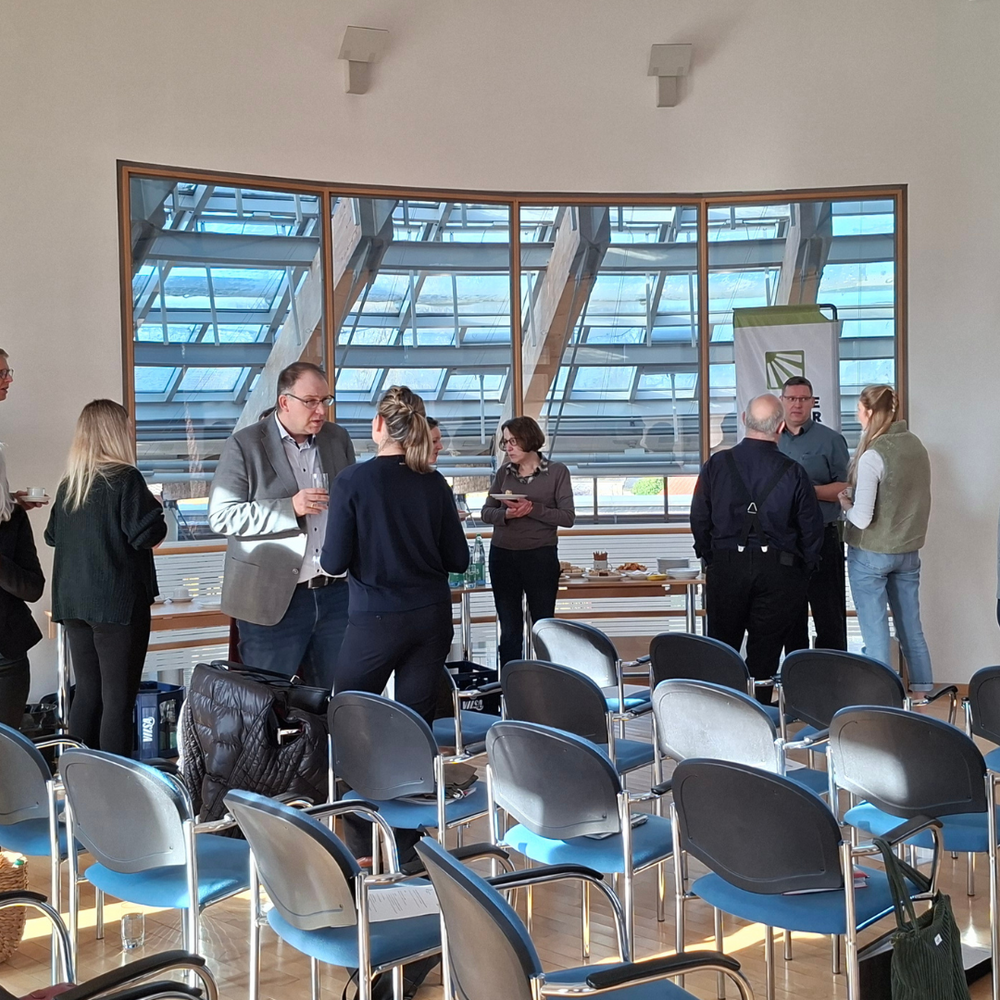 | Ⓒ Deltaland Mehrere Personen stehen in kleinen Gruppen in einem hellen Seminarraum und unterhalten sich während einer Pause; im Vordergrund sind leere Reihenstühle zu sehen. Rechts gestikuliert eine Frau neben einer Leinwand, während links andere Teilnehmende Getränke und Snacks vom Tisch nehmen.