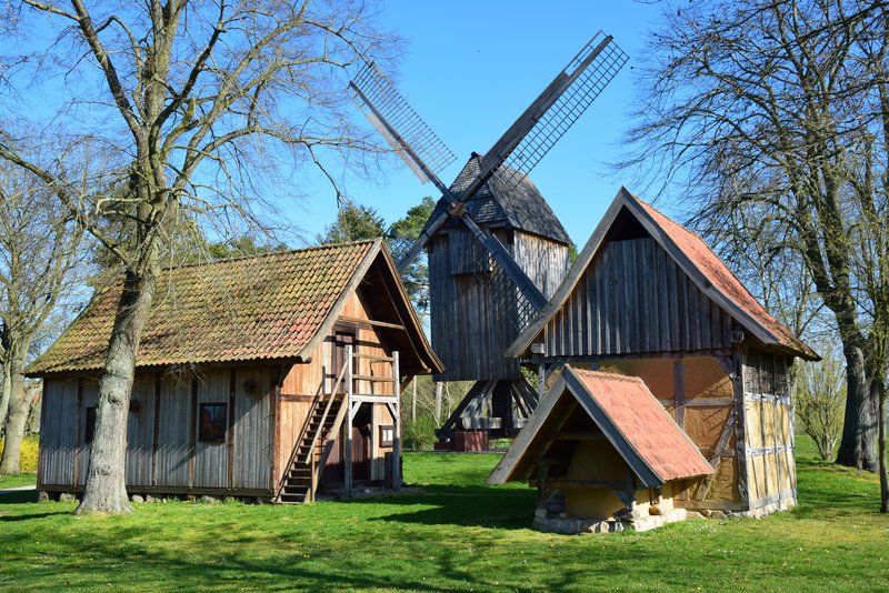 Das Bild zeigt eine traditionelle Dorfszene mit mehreren historischen Holzgebäuden und einer markanten Windmühle. Die idyllische Landschaft mit Bäumen und grünen Rasenflächen verleiht der Szene einen idyllischen, ländlichen Charakter.