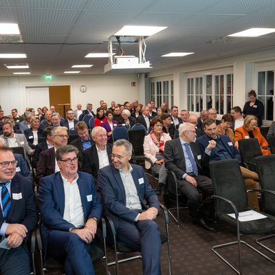 Auf dem Bild ist eine Gruppe von Personen zu sehen, die in einem Innenraum auf Stühlen sitzen. Die Personen sind  formell gekleidet und scheinen an einer Veranstaltung teilzunehmen. 