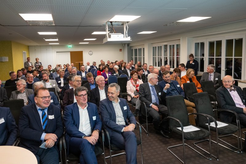 Auf dem Bild ist eine Gruppe von Personen zu sehen, die in einem Innenraum auf Stühlen sitzen. Die Personen sind  formell gekleidet und scheinen an einer Veranstaltung teilzunehmen. 