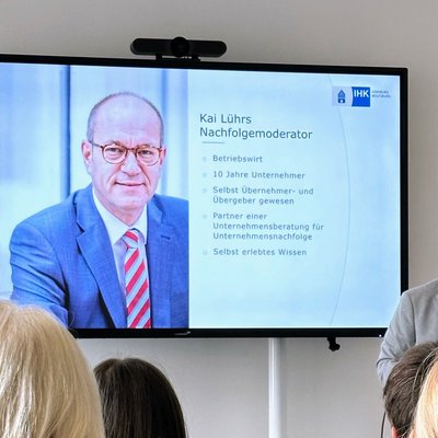 IHK-Nachfolgemoderator Kai Lührs (Mann mit Brille in hellgrauem Sakko) hält einen Vortrag. Neben im ist ein großer Monitor zu sehen, der die aktuelle Vortragsfolie von Kai Lührs darstellt (hier ein paar Infos zur Person Lührs selbst).