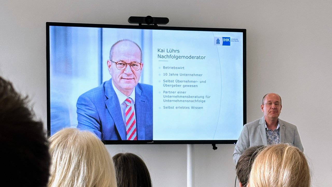 IHK-Nachfolgemoderator Kai Lührs (Mann mit Brille in hellgrauem Sakko) hält einen Vortrag. Neben im ist ein großer Monitor zu sehen, der die aktuelle Vortragsfolie von Kai Lührs darstellt (hier ein paar Infos zur Person Lührs selbst).