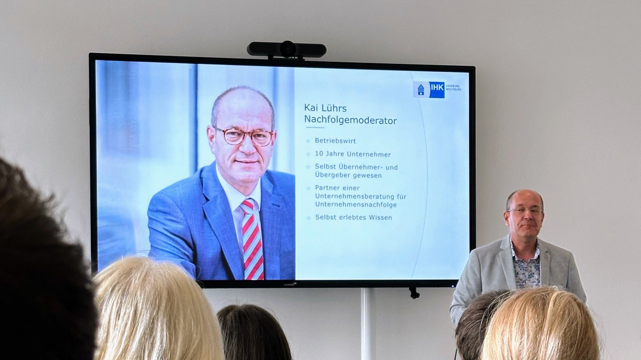 IHK-Nachfolgemoderator Kai Lührs (Mann mit Brille in hellgrauem Sakko) hält einen Vortrag. Neben im ist ein großer Monitor zu sehen, der die aktuelle Vortragsfolie von Kai Lührs darstellt (hier ein paar Infos zur Person Lührs selbst).