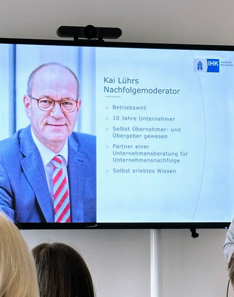 IHK-Nachfolgemoderator Kai Lührs (Mann mit Brille in hellgrauem Sakko) hält einen Vortrag. Neben im ist ein großer Monitor zu sehen, der die aktuelle Vortragsfolie von Kai Lührs darstellt (hier ein paar Infos zur Person Lührs selbst).