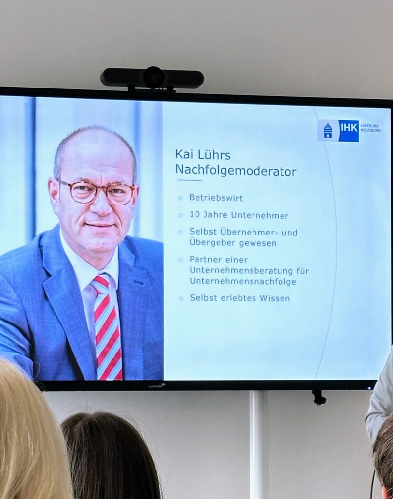 IHK-Nachfolgemoderator Kai Lührs (Mann mit Brille in hellgrauem Sakko) hält einen Vortrag. Neben im ist ein großer Monitor zu sehen, der die aktuelle Vortragsfolie von Kai Lührs darstellt (hier ein paar Infos zur Person Lührs selbst).