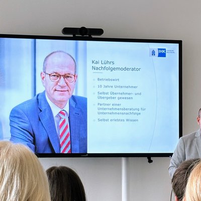 IHK-Nachfolgemoderator Kai Lührs (Mann mit Brille in hellgrauem Sakko) hält einen Vortrag. Neben im ist ein großer Monitor zu sehen, der die aktuelle Vortragsfolie von Kai Lührs darstellt (hier ein paar Infos zur Person Lührs selbst).